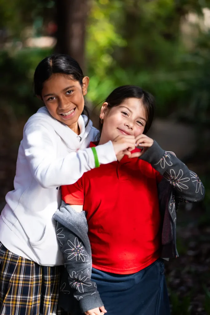 Two smiling children outdoors making heart gesture.